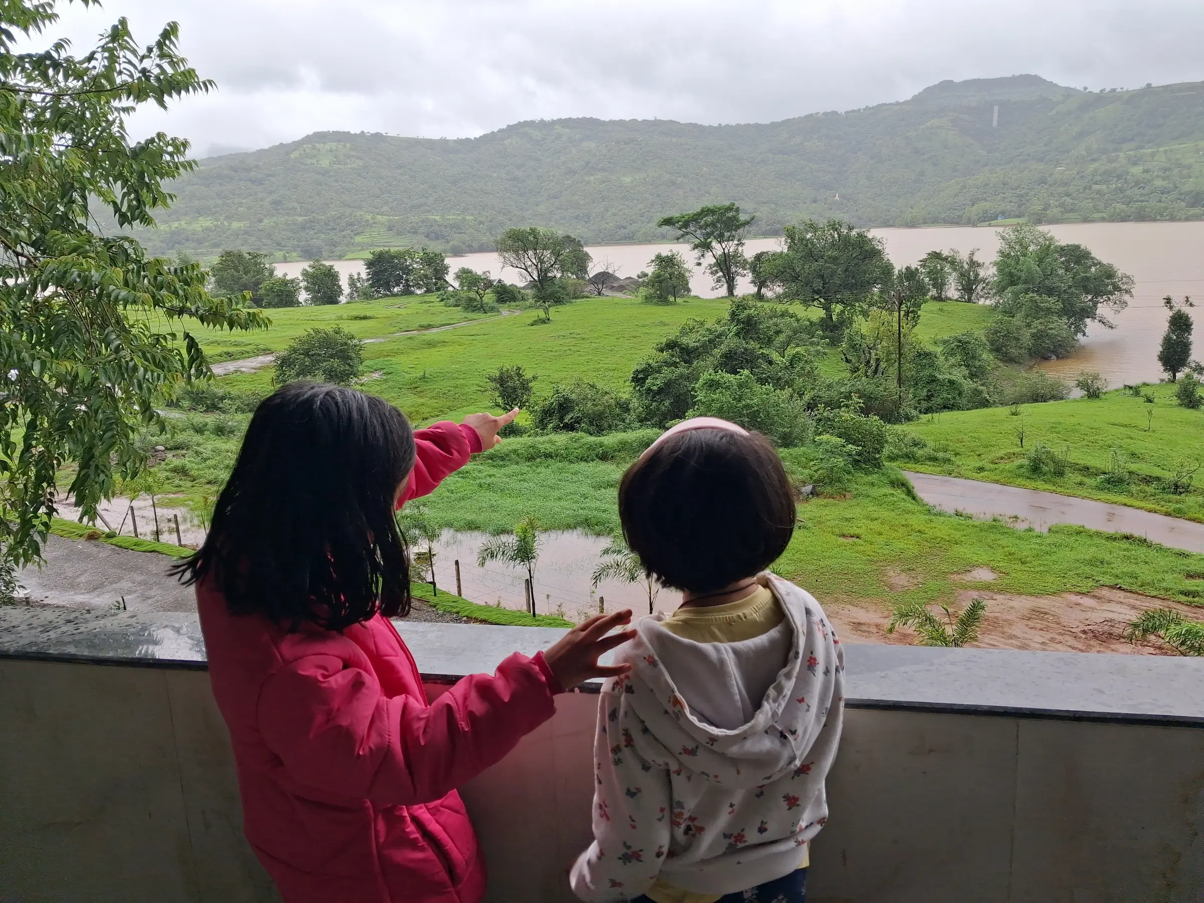 Two children admire a lush green landscape with hills and a river.